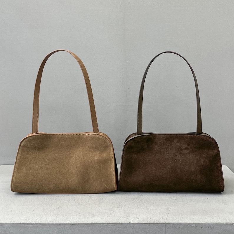 May include: Two suede shoulder bags in the image. One bag is a light tan color, and the other is a dark brown. Both bags have a rectangular shape with rounded corners and long, thin handles. The bags are displayed on a light gray surface.