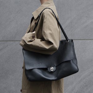 Puede incluir: Un bolso de hombro grande de cuero negro con textura granulada, que presenta una solapa ancha y un cierre de giro de metal plateado. El bolso tiene dos correas de hombro de cuero negro finas unidas con anillas de metal plateado.