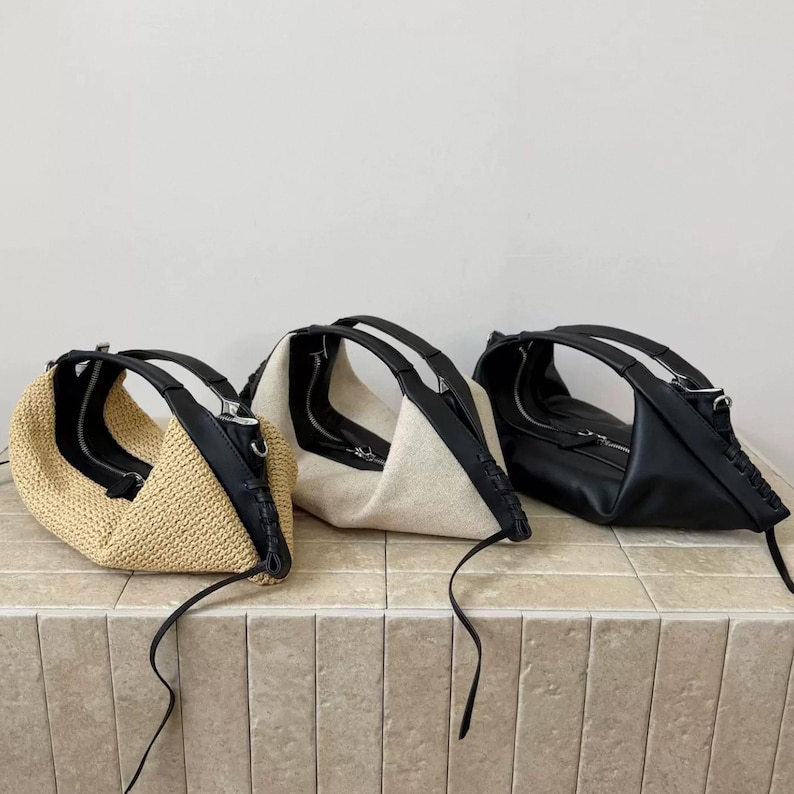 May include: Three crescent-shaped handbags arranged in a row. The first is made of tan woven material, the second of off-white fabric, and the third of smooth black material. Each bag features a black leather-like handle and a silver zipper closure.