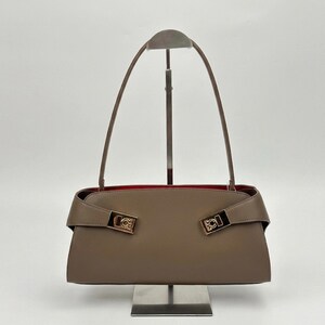 May include: A taupe-colored handbag with a rectangular shape and a red interior. The bag features two gold-colored rectangular clasps and long, thin handles. The bag is displayed on a metal stand against a white background.