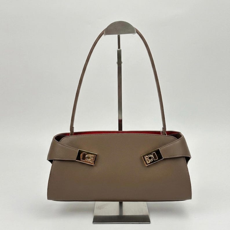 May include: A taupe-colored handbag with a rectangular shape and a red interior. The bag features two gold-colored rectangular clasps and long, thin handles. The bag is displayed on a metal stand against a white background.