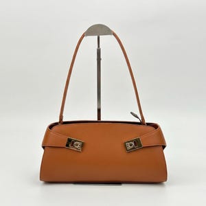 May include: A brown leather handbag with a rectangular shape and a top handle. The bag features gold-colored hardware on the front and a flap closure. The bag is displayed on a metal stand against a white background.