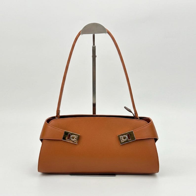 May include: A brown leather handbag with a rectangular shape and a top handle. The bag features gold-colored hardware on the front and a flap closure. The bag is displayed on a metal stand against a white background.