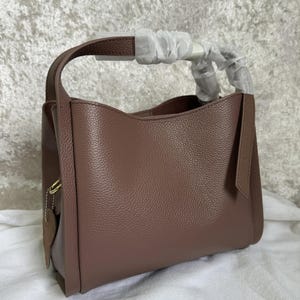 May include: A mauve leather tote bag with a single top handle and a zipper closure. The bag has a textured pebbled leather finish.