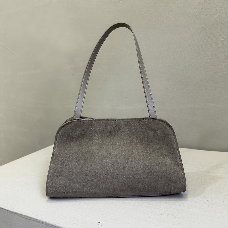 May include: A gray suede shoulder bag with a rectangular body and a curved top. The bag has a single, thin strap and a minimalist design. The bag is sitting on a white surface.