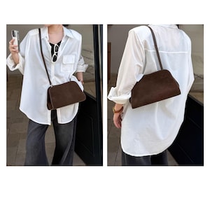 May include: A brown suede crossbody bag with a long strap. The bag has a rectangular shape with rounded edges. The bag is worn over a white button-down shirt and dark pants.