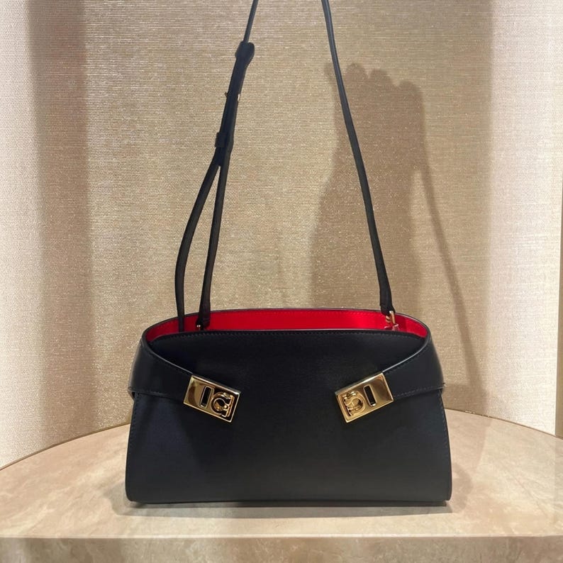 May include: A black handbag with a red interior and gold-colored hardware. The bag has a rectangular shape with a flap closure and a long, thin strap. The gold hardware is rectangular with a decorative design.