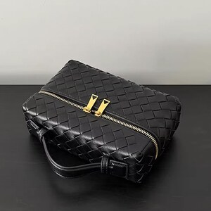 Puede incluir: Bolso de mano de cuero negro tejido con cremallera y asa doradas. El bolso tiene forma rectangular y un patr&oacute;n tejido. La cremallera tiene dos tiradores dorados. El bolso est&aacute; sobre una superficie negra.