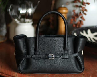May include: A black handbag with a textured surface, featuring two rounded top handles and a horizontal strap across the front secured by a silver-toned metal buckle. The bag has a structured base and slightly gathered sides, creating a compact shape.