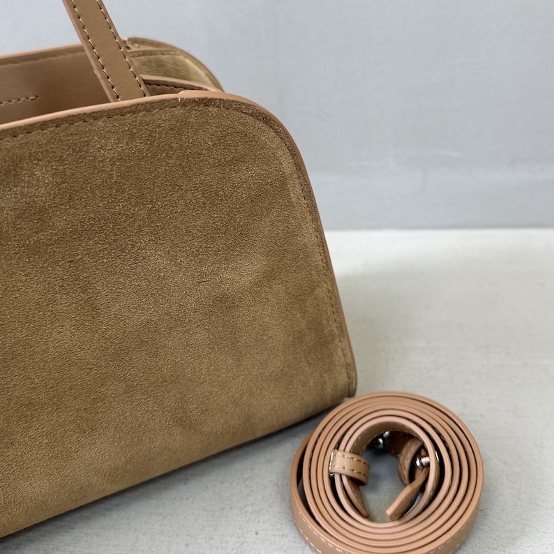 May include: A tan suede handbag with a matching shoulder strap. The bag has a rounded top and a structured design. The strap is coiled and ready for use. The bag's handle is visible.