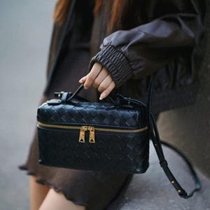 Puede incluir: Bolso negro de cuero tejido con cremallera y herrajes dorados. El bolso tiene un asa superior y una correa para el hombro desmontable. El bolso lo sostiene una persona que lleva una chaqueta marr&oacute;n.