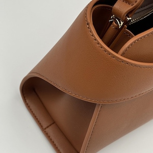 May include: Close-up of a brown leather handbag with a curved top and a rectangular base. The bag features a zipper closure and a handle. The stitching is visible along the edges, and the leather has a smooth texture.