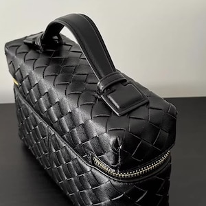 Puede incluir: Bolso de mano de cuero negro con dise&ntilde;o tejido. El bolso tiene un asa superior, una cremallera dorada y una forma rectangular. El asa est&aacute; asegurada con una hebilla.