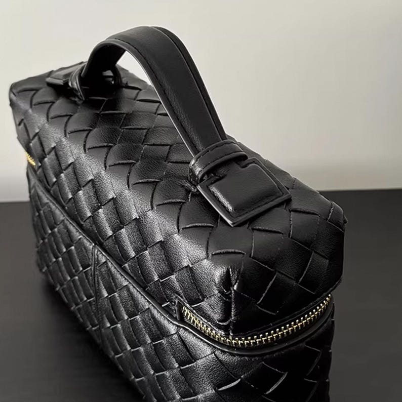 Puede incluir: Bolso de mano de cuero negro con dise&ntilde;o tejido. El bolso tiene un asa superior, una cremallera dorada y una forma rectangular. El asa est&aacute; asegurada con una hebilla.