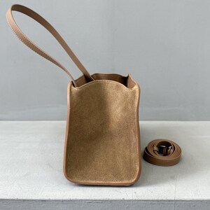 May include: A tan-colored handbag with a long handle and a detachable shoulder strap. The bag has a rectangular shape with rounded corners and a suede-like texture. The handle and trim are a lighter shade of tan.