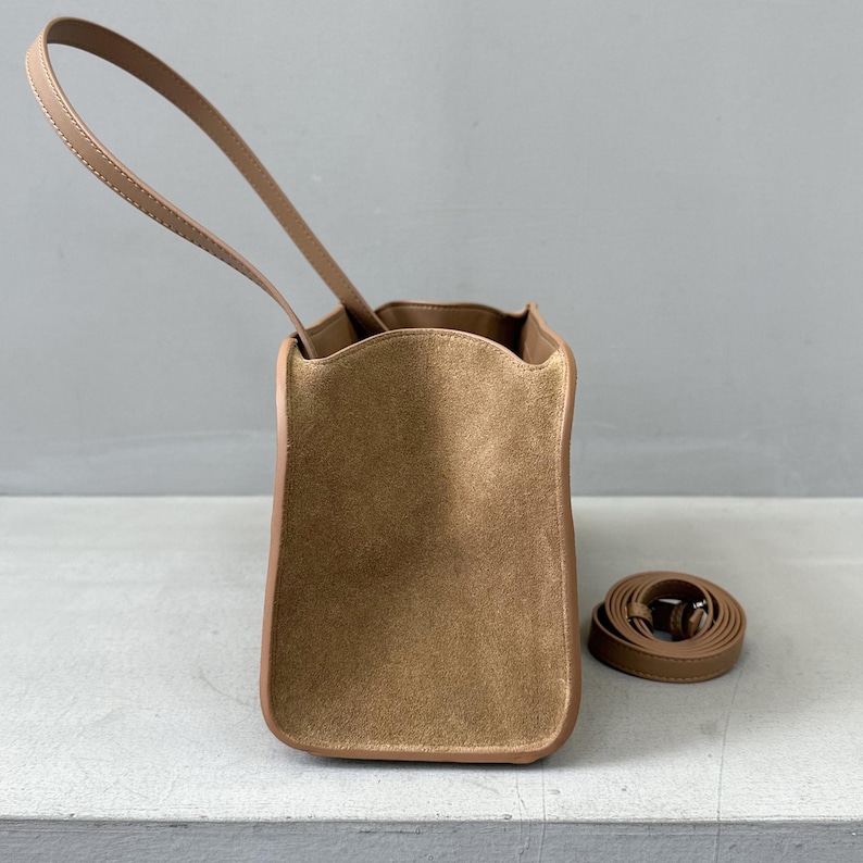 May include: A tan-colored handbag with a long handle and a detachable shoulder strap. The bag has a rectangular shape with rounded corners and a suede-like texture. The handle and trim are a lighter shade of tan.