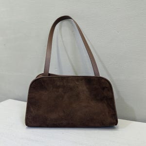 May include: A brown suede shoulder bag with a rounded rectangular shape. The bag has a matching brown handle and a light brown strap. The bag is sitting on a white surface.