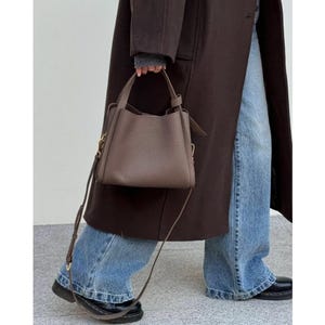 May include: A brown leather tote bag with a long strap. The bag has a top handle and a flap closure. The bag is being carried by a person wearing a brown coat and blue jeans.