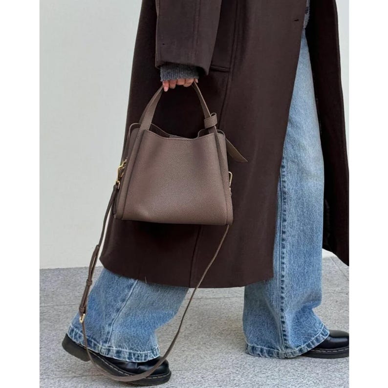 May include: A brown leather tote bag with a long strap. The bag has a top handle and a flap closure. The bag is being carried by a person wearing a brown coat and blue jeans.