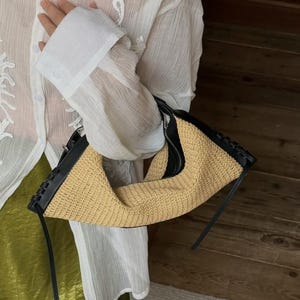 May include: A crescent-shaped handbag featuring a woven tan straw body with black leather trim and edges. The bag includes a black zipper closure along the top and black leather side panels with a decorative braided stitch detail along the seams.