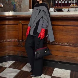 May include: A black shoulder bag with a gold clasp and a burgundy interior is held by a person. The person is wearing a gray and black sweater, black pants, and red gloves. The background features a wooden bar and a checkered floor.