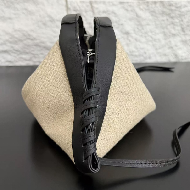 May include: A triangular-shaped handbag featuring beige canvas side panels and a black leather-like centre strip. The centre strip includes a decorative black lace-up detail, a silver-toned metal zip closure, and a thin black shoulder strap.