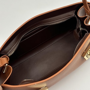May include: Open view of a brown leather handbag. The interior features a dark brown lining, a zippered pocket, and a small pocket. The bag has a light brown exterior with gold-colored hardware and a top handle.