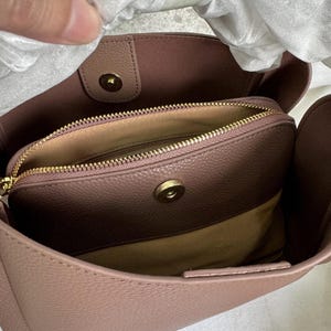 May include: A mauve-colored handbag with a gold zipper and a gold button closure. The bag has a soft, textured fabric lining.