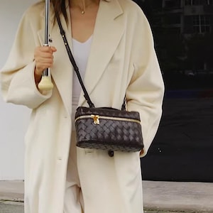 Puede incluir: Bolso bandolera de cuero tejido marr&oacute;n oscuro con cremallera y herrajes dorados. El bolso rectangular tiene una correa larga y fina. El bolso se lleva sobre un abrigo color crema.