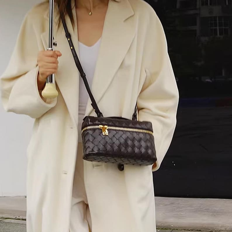 Puede incluir: Bolso bandolera de cuero tejido marr&oacute;n oscuro con cremallera y herrajes dorados. El bolso rectangular tiene una correa larga y fina. El bolso se lleva sobre un abrigo color crema.