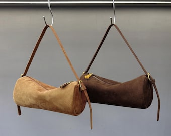 May include: Two suede barrel-shaped handbags in shades of brown hang from metal hooks. Each bag features a long, thin strap and a zipper closure with gold-toned hardware. The bags are displayed against a neutral background.