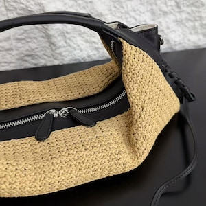 May include: A handbag featuring a combination of black leather and beige woven straw-like material. The bag has a curved top handle, a silver-toned metal zip closure with two black leather pull tabs, and a structured design with visible stitching.