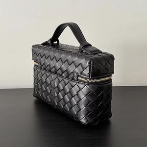 Puede incluir: Bolso de mano de cuero negro tejido con asa superior y cremallera dorada. El bolso rectangular presenta un dise&ntilde;o de tejido de cesta y un asa peque&ntilde;a para llevarlo. El bolso est&aacute; sobre una superficie negra.