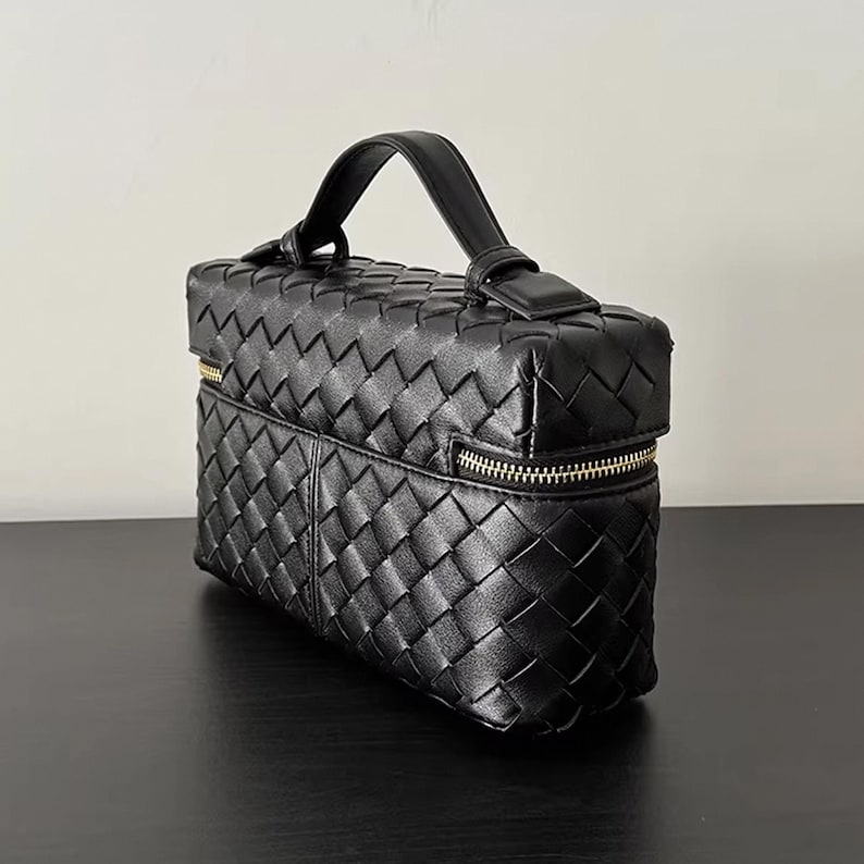 Puede incluir: Bolso de mano de cuero negro tejido con asa superior y cremallera dorada. El bolso rectangular presenta un dise&ntilde;o de tejido de cesta y un asa peque&ntilde;a para llevarlo. El bolso est&aacute; sobre una superficie negra.