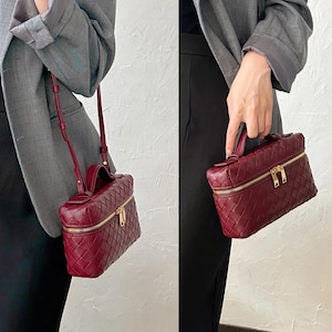 Puede incluir: Un bolso de mano de cuero tejido de color burdeos con forma rectangular. El bolso tiene un asa superior, una correa de hombro desmontable y un cierre de cremallera dorado. El bolso se muestra en dos vistas diferentes.