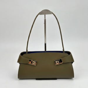 May include: Olive green handbag with a rectangular shape and a blue interior. The bag features two gold-colored clasps and a long, thin handle. The bag is displayed on a silver stand against a white background.