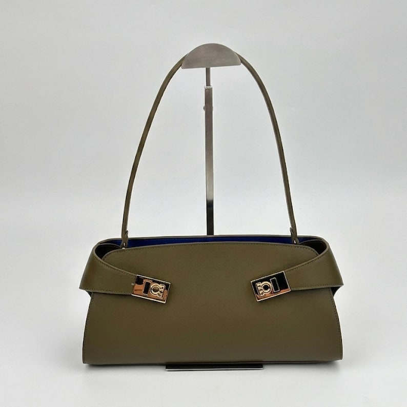 May include: Olive green handbag with a rectangular shape and a blue interior. The bag features two gold-colored clasps and a long, thin handle. The bag is displayed on a silver stand against a white background.