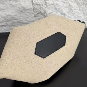 May include: A beige canvas pouch featuring a textured, woven fabric construction. The pouch is accented with a centred, black, hexagonal-shaped leather patch that is stitched onto the front surface, providing a contrast to the light material.
