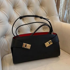May include: Black handbag with a red interior. The bag has a rectangular shape with a flap closure and gold-colored hardware. It features a top handle and a shoulder strap. The bag is sitting on a white chair.