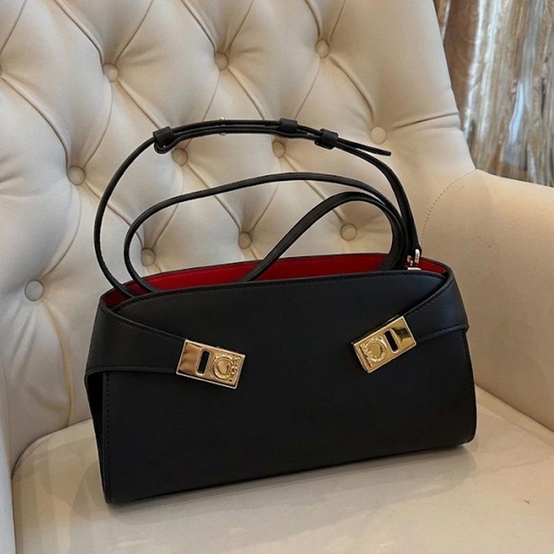 May include: Black handbag with a red interior. The bag has a rectangular shape with a flap closure and gold-colored hardware. It features a top handle and a shoulder strap. The bag is sitting on a white chair.