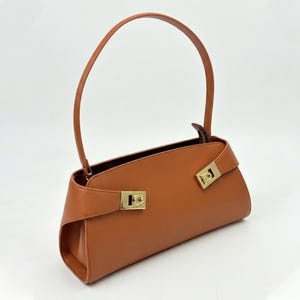 May include: A tan leather handbag with a curved handle and gold-colored hardware. The bag has a rectangular shape with a flap closure and a zipper. The bag is made of smooth leather and has a minimalist design.