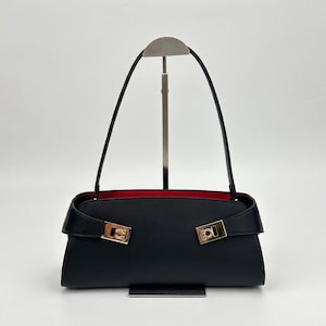 May include: Black handbag with a rectangular shape and a red interior. The bag features a flap closure with gold-colored hardware and long black straps. The bag is displayed on a silver stand against a white background.