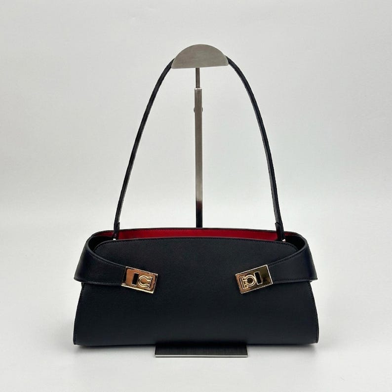 May include: Black handbag with a rectangular shape and a red interior. The bag features a flap closure with gold-colored hardware and long black straps. The bag is displayed on a silver stand against a white background.