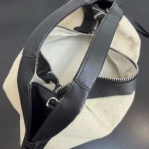 May include: A handbag featuring a beige canvas body with black leather trim and handles. The bag has a top zip closure with silver-toned hardware, including a metal zip pull and a small D-ring attachment on the side of the black leather strap.