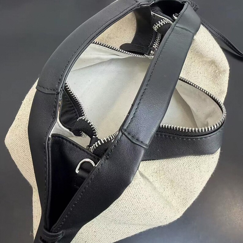 May include: A handbag featuring a beige canvas body with black leather trim and handles. The bag has a top zip closure with silver-toned hardware, including a metal zip pull and a small D-ring attachment on the side of the black leather strap.