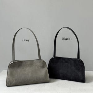 May include: Two suede shoulder bags, one gray and one black, are displayed against a neutral background. Each bag has a rectangular body with a curved top and a matching handle. The word "Gray" is above the gray bag, and "Black" is above the black bag.