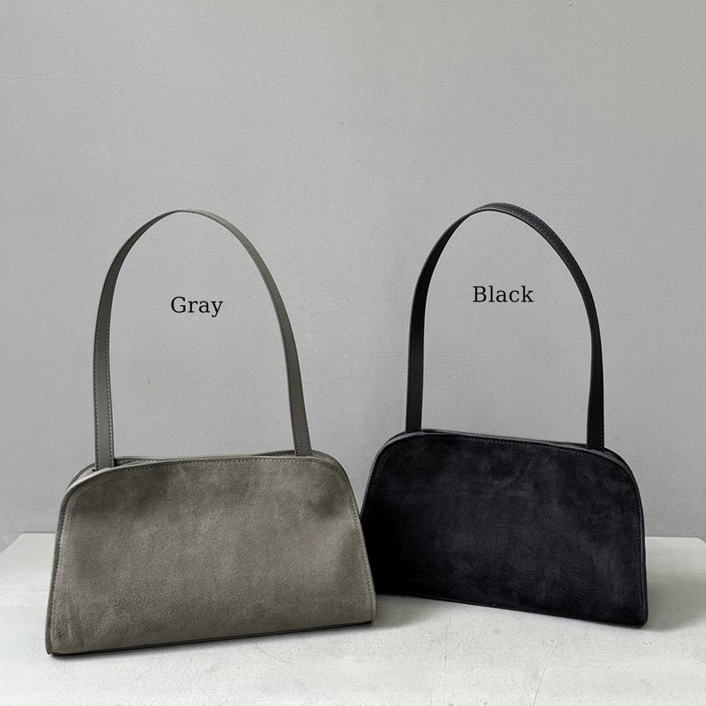May include: Two suede shoulder bags, one gray and one black, are displayed against a neutral background. Each bag has a rectangular body with a curved top and a matching handle. The word "Gray" is above the gray bag, and "Black" is above the black bag.