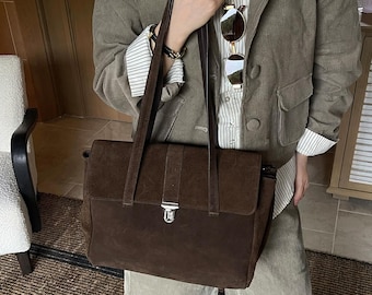 May include: A dark brown suede tote bag featuring a rectangular flap closure, two long shoulder straps, and a central silver-toned metal push-lock clasp. The bag has a structured silhouette with a smooth, matte texture and clean, minimalist lines.