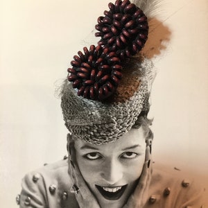 May include: A black and white photo of a woman wearing a hat with two large, dark red, circular, decorative embellishments. The woman is smiling and has her hands on her cheeks.