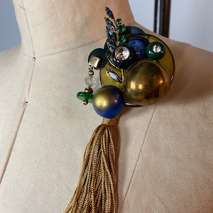 May include: A gold, blue, and green statement brooch with a tassel. The brooch features a variety of textures and materials, including glass, metal, and fabric.